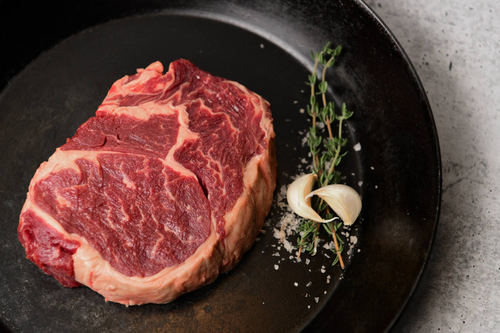 A raw ribeye steak on on a dark plate, with two cloves of garlic and sprigs of thyme.