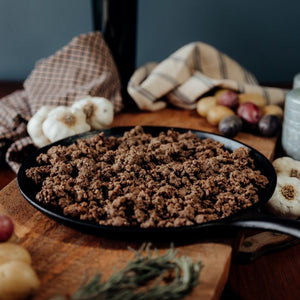 A black skillet with cooked 75% lean ground beef in a paper wrap on a wooden cutting board with vegetables and a plaid cloth in the background