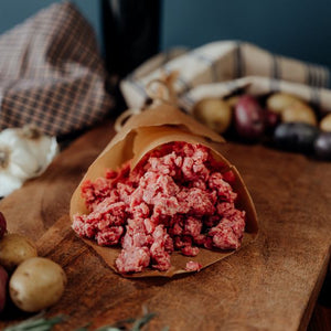Raw 75% lean ground beef in a paper wrap on a wooden cutting board with vegetables and a plaid cloth in the background.