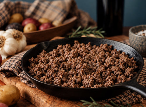 Cooked grassfed 90% Lean ground beef in a black cast iron skillet, styled on a wooden cutting board with garlic, potatoes, herbs, and kitchen props in a rustic kitchen setting.