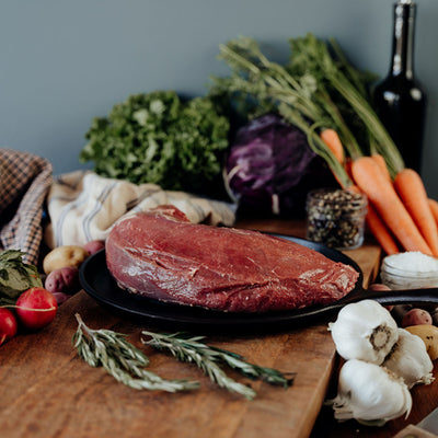 Raw beef tenderloin on a cutting board with a variety of vegetables on a wooden table