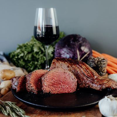 A cooked and sliced beef tenderloin on a cutting board with a variety of vegetables and a glass on a wooden table