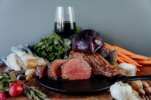 A cooked and sliced beef tenderloin on a cutting board with a variety of vegetables and a glass on a wooden table