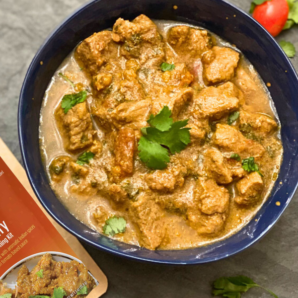 Bowl of curry with chunks of meat and herbs, next to a packet of seasoning mix on a gray surface.