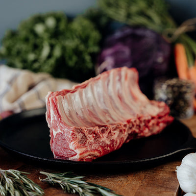 Raw Rack of Lamb Split Loin Roast on a black plate with vegetables in the background