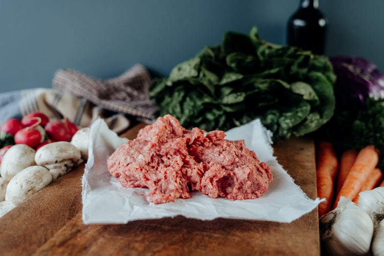 Ground meat on a wooden cutting board with vegetables, garlic, and a bottle in the background