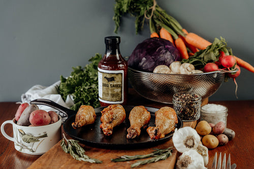 Cooked Chicken Drumsticks seasoned with BBQ Sauce on a black skillet with vegetables and a bottle of BBQ Sauce in the background.