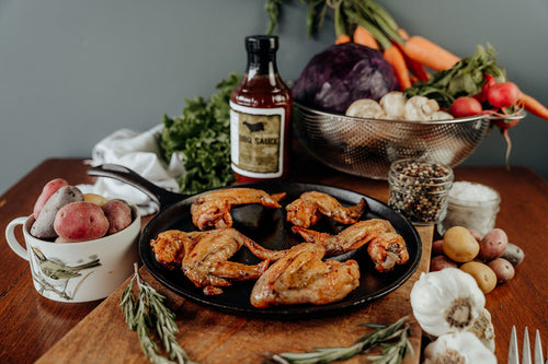 On a black plate five cooked Pasture-Raised Chicken Wings seasoned with US Wellness Meats BBQ Sauce sits on a wooden cutting board with vegetables, seasonings, a bottle of BBQ Sauce, and kitchen utensils on a table.