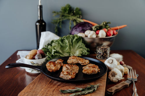 Four cooked Boneless, Skinless Chicken Thighs on a wooden cutting board with vegetables, spices, and a bottle in the background.