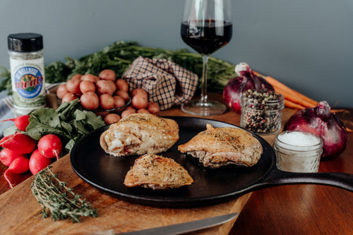 Raw bone in chicken thighs, pasture-raised chicken on a black plate with vegetables, a bottle, and spices on a wooden board.