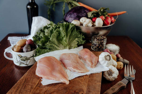 Raw Pasture-Raised Chicken Breast on a wooden cutting board with vegetables and a knife in the background.
