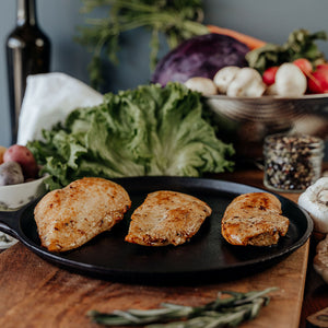 Three raw Pasture-Raised Chicken Breast on a wooden cutting board with vegetables and a knife in the background.