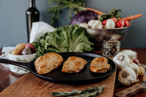 Three raw Pasture-Raised Chicken Breast on a wooden cutting board with vegetables and a knife in the background.