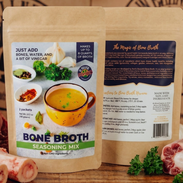 Two stand-up pouches of Bone Broth Seasoning Mix are shown front and back on a wooden surface, surrounded by raw bones, garlic, parsley, and other cooking ingredients. The background is a branded brown paper backdrop.
