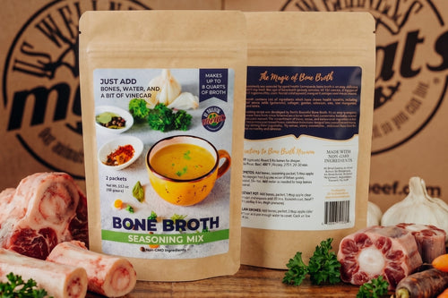 Two stand-up pouches of Bone Broth Seasoning Mix are shown front and back on a wooden surface, surrounded by raw bones, garlic, parsley, and other cooking ingredients. The background is a branded brown paper backdrop.