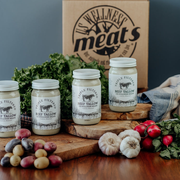 Four jars of triple filtered beef tallow on a wooden surface with a 'Wellness Meats' box, cloths, and vegetables in the background.