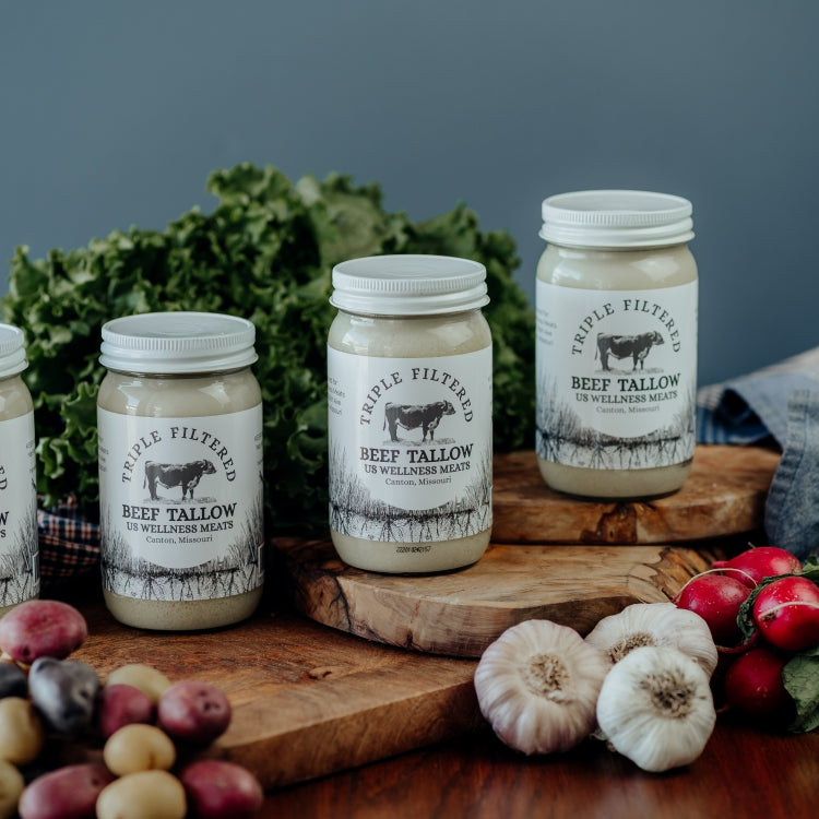 Four jars of triple filtered beef tallow on a wooden surface with cloths and vegetables in the background.