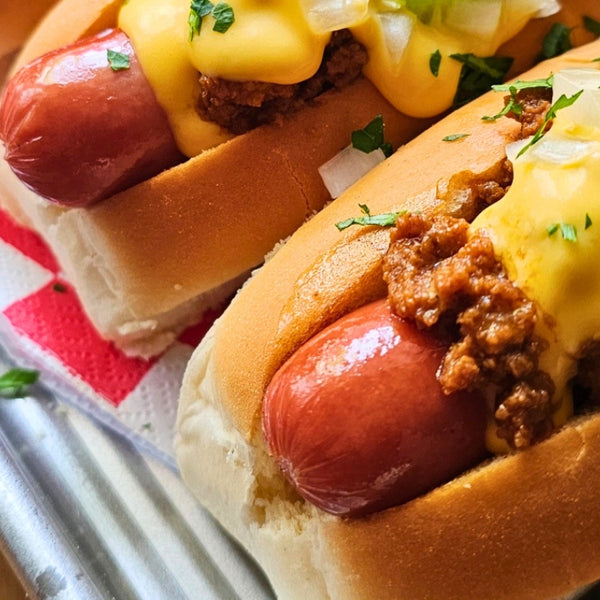 Two chili dog hot dogs topped with melted cheese, ground chili, onions, and jalapeño slices on a tray with a red-and-white checkered paper liner.