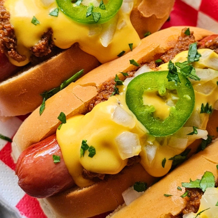 Three loaded beef franks in buns topped with melted yellow cheese, jalapeño slices, onions, and chopped green herbs, served on a red-and-white checkered paper liner.