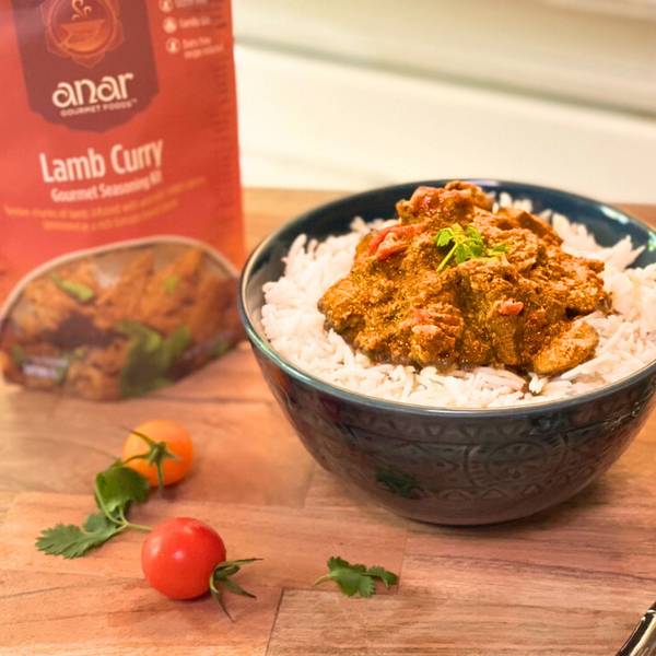 Bowl of lamb curry with rice next to a box of anar Lamb Curry seasoning on a wooden surface.