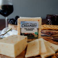 One block of Raw Jalapeno Cheese with slices on a wooden block in front of a labeled Lykens Valley Creamery Raw Jalapeno Cheese block with grapes, crackers, berries, jam, and a wine glass in the back