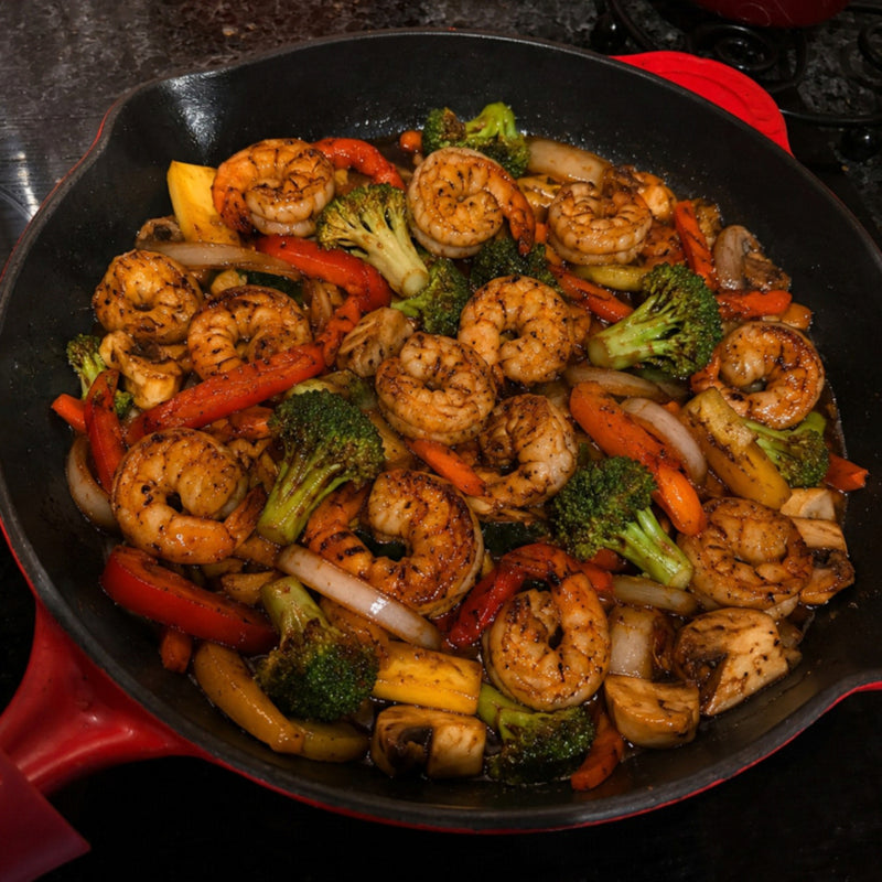 A close-up shot of a shrimp and vegetable stir-fry in a black cast iron skillet with a red handle, set against a dark kitchen background.