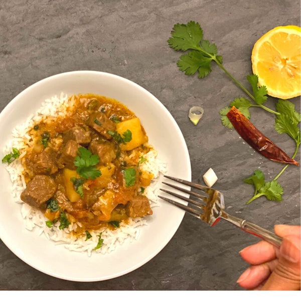 Curry dish with rice on a white plate, garnished with lemon, cilantro, and a red chili pepper, on a dark surface.