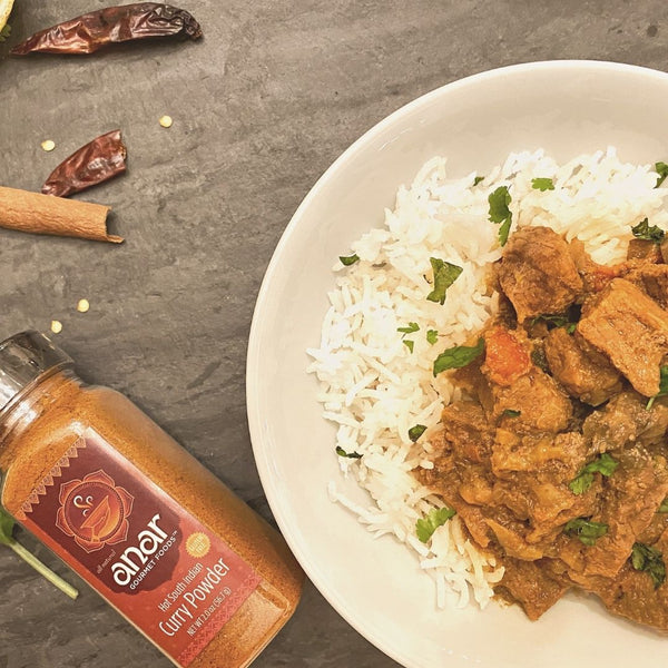 Curry dish with rice and spices on a gray surface, featuring a bottle of 'anar' curry powder.