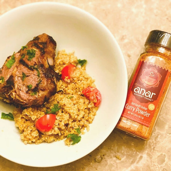 Bowl of quinoa with grilled meat and tomatoes next to a bottle of anar curry powder.
