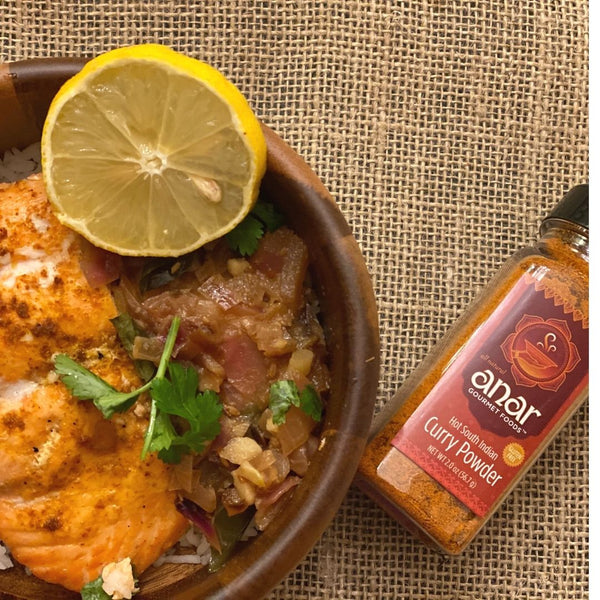 Bowl of curry-seasoned white fish over rice topped with sautéed onions, cilantro, and a lemon slice, next to a jar of Anar Gourmet Foods Hot South Indian Curry Powder on a burlap background.
