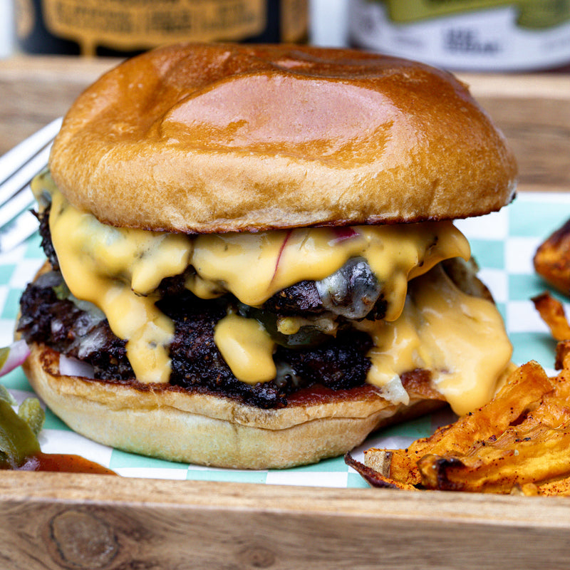 A close-up of a gourmet burger with melted yellow cheese, served on a wooden board with potato chips and a blurred bottle in the background.
