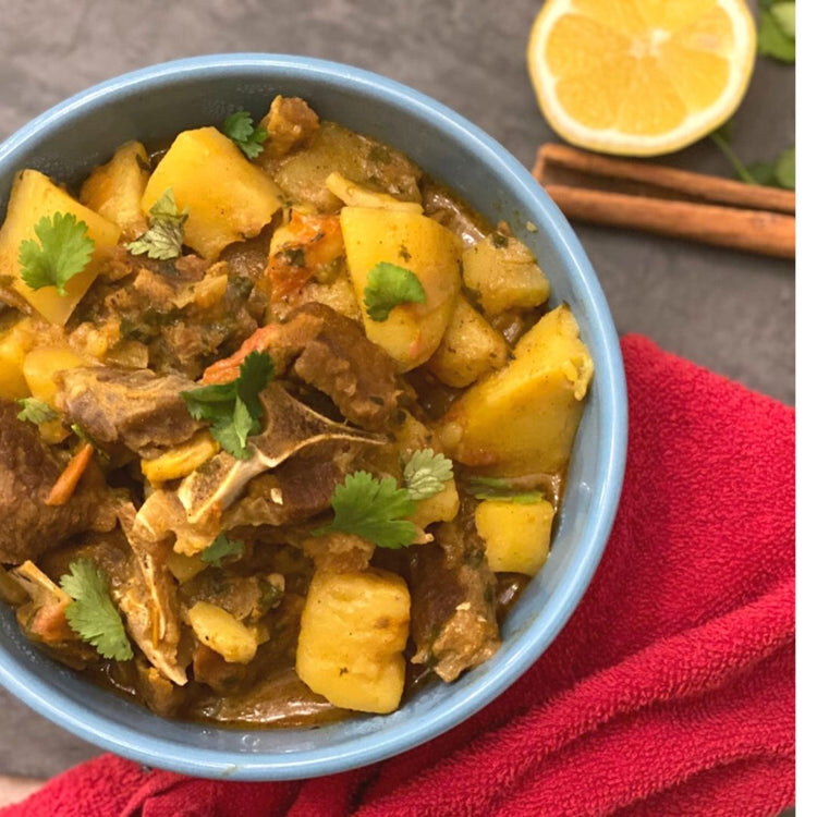 Bowl of curry with potatoes and meat on a red cloth with a lemon and cinnamon sticks in the background.