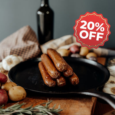 grassfed beef franks on a pan with a 20% off sign, surrounded by vegetables and a bottle, on a wooden surface.