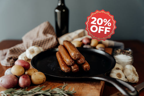 grassfed beef franks on a pan with a 20% off sign, surrounded by vegetables and a bottle, on a wooden surface.