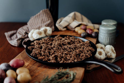 A black skillet on a cooked 85% lean ground beef on a wooden cutting board with vegetables, cloth, and a bottle in the background.