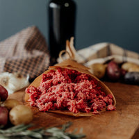 Raw 85%lean ground beef in a paper wrap on a wooden cutting board with vegetables, cloth, and a bottle in the background.