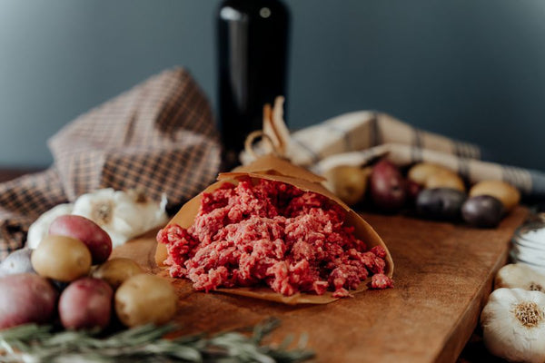 Raw 85%lean ground beef in a paper wrap on a wooden cutting board with vegetables, cloth, and a bottle in the background.