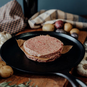 Stack of two raw 55 lean beef ground patties on a black plate with garlic and potatoes in the background.