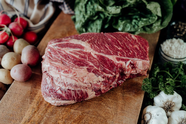 Raw grassfed beef chuck roast on a wooden cutting board with a variety of vegetables in the background