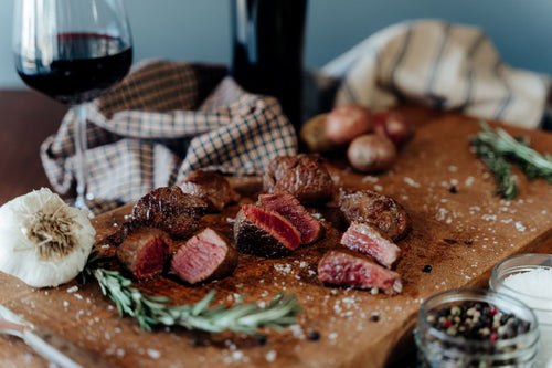 Wooden cutting board with grilled and sliced Grassfed Beef Tenderloin Kabobs, garlic, rosemary, and a glass of red wine.