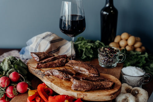 A grilled and sliced grassfed beef heart on a black plate with a variety of vegetables, herbs, and a glass of wine on a wooden surface