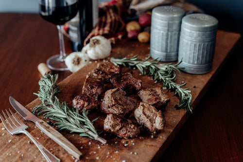 Grilled Grassfed Beef NY Strip Kabobs on a wooden cutting board with spices, potatoes, cloth, and a glass of wine in the background