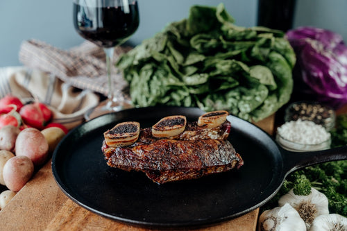 A smoked grassfed beef bone-in short rib on a black plate with a variety of vegetables in the background