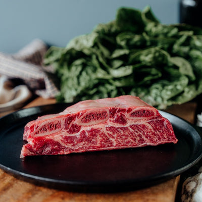 A raw grassfed beef bone-in short rib on a black plate with a variety of vegetables in the background