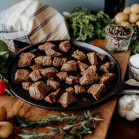 Cooked Grass-Fed Stew Beef in a black skillet surrounded by vegetables on a wooden surface