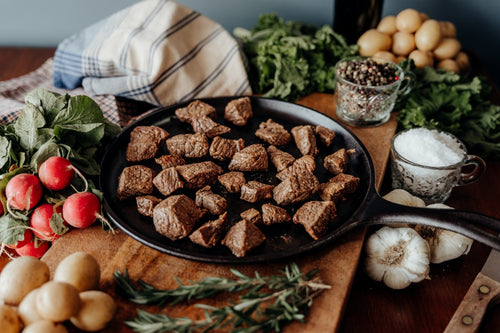 Cooked Grass-Fed Stew Beef in a black skillet surrounded by vegetables on a wooden surface
