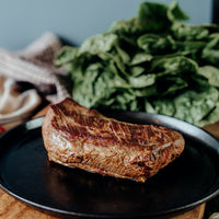 Sitting on a black skillet is a Grilled grassfed beef tri-tip roast on a wooden cutting board with vegetables and herbs.