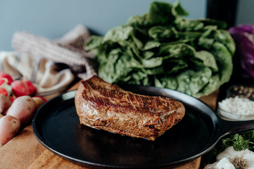 Sitting on a black skillet is a Grilled grassfed beef tri-tip roast on a wooden cutting board with vegetables and herbs.