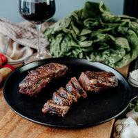 Two pieces of grilled and sliced Boneless Beef Short Ribs on a black plate with a variety of vegetables in the background.