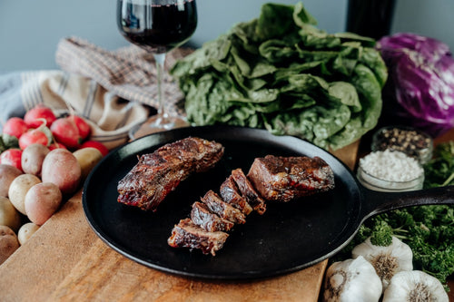 Two pieces of grilled and sliced Boneless Beef Short Ribs on a black plate with a variety of vegetables in the background.